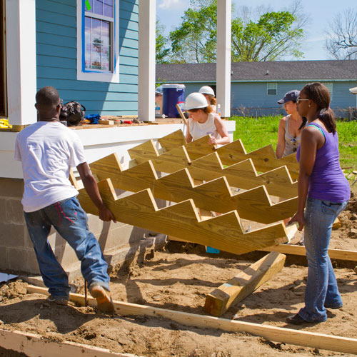 Four students collaborate to lift a wooden staircase frame in front of a blue house under construction. The scene is energetic and sunny, suggesting teamwork.