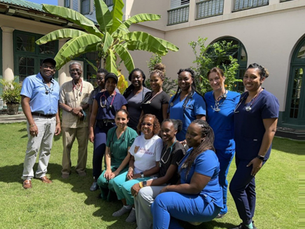 A group of medical professionals, mostly women in blue or green scrubs, stand and kneel on a grassy lawn in front of a building.