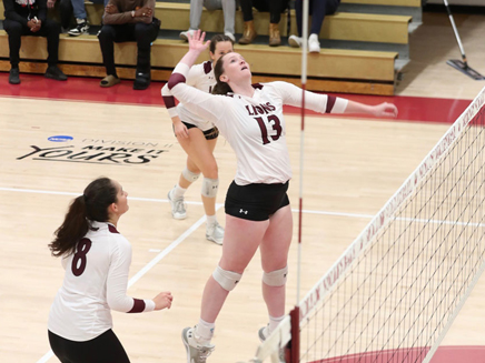 A female volleyball player in a white jersey with the number 13 and the word "LIONS" on it jumps to hit the ball during a game. Another female player in a white jersey with the number 8 is looking up at the ball.