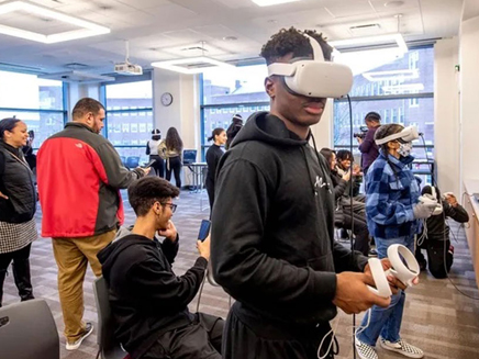 Several people are using virtual reality headsets in a bright room. A young man in the foreground is holding VR controllers.