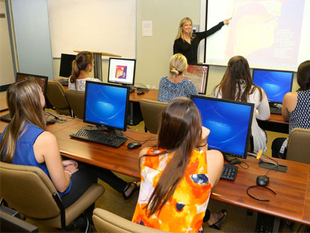 A female instructor points to a diagram on a projector screen while several female students sit at computers in a classroom.