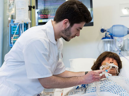 Male Molloy nurssing student adjusts the breathing tube of a patient in a hospital bed, with medical equipment in the background.