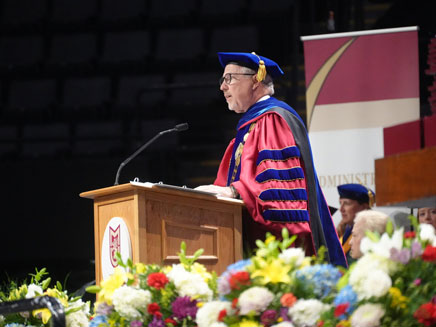 President James Lentini speaks at a podium at Molloy University Commencement, surrounded by colorful flowers.