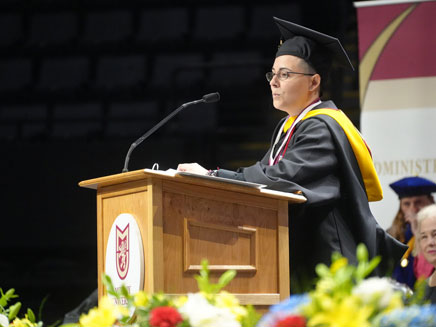 Valedictorian, Victoria Franco, in cap and gown speaks at a wooden podium during commencement ceremony.