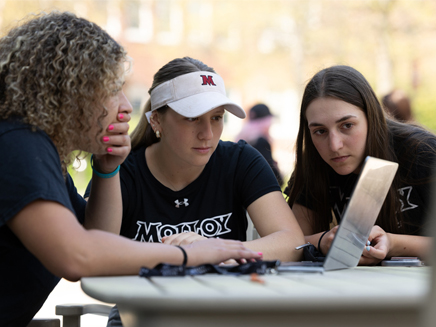 Three young women sit at an outdoor table closely looking at a laptop screen together.
