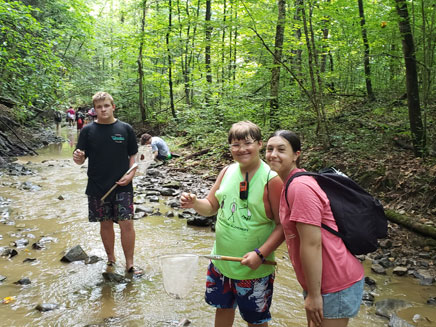 Two young people stand in a shallow stream surrounded by trees. They each hold a net, seemingly participating in a nature activity.