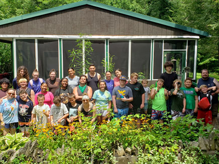 A large group of people, including adults and children, are gathered for a photo in front of a rustic cabin in the Appalachias