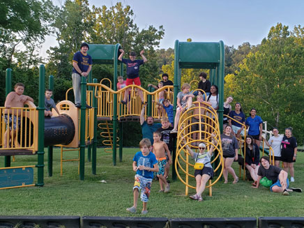 A large group of people, mostly children and teenagers, are posing on and around a playground structure. The group is smiling and appears to be having fun together.