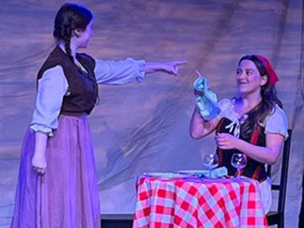 Two Cap21 stduents in theatrical costumes are on stage. One woman is pointing at the other woman, who is seated at a table covered with a red and white checkered tablecloth.