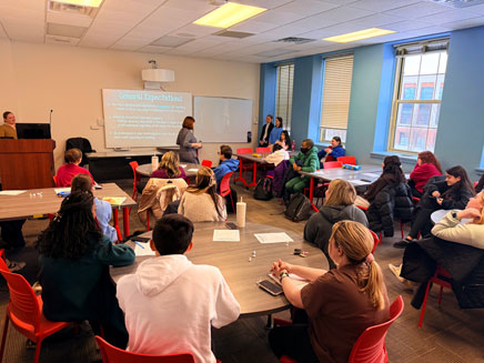 A classroom setting where a presenter is addressing a group of students seated at tables.