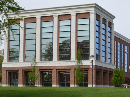 Side view of the Barbara H. Hagan School of Nursing on the Molloy University Campus. A large brick and glass building with tall windows and classical columns stands surrounded by green grass and trees, under a clear sky.
