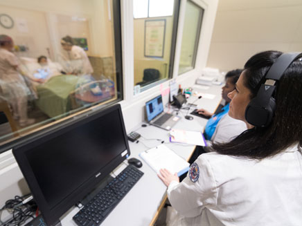 A healthcare professional wearing headphones monitors a simulation room through a window. Two individuals assist a patient on a bed inside.