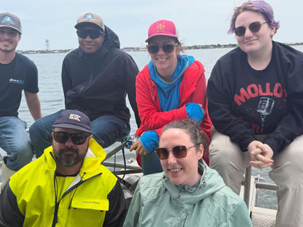 Dr. Elizabeth (Liz) Suter and a group of people on a boat, smiling under a cloudy sky. The sea and distant shoreline are visible.